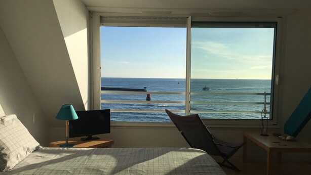 A bedroom with a large window offering a view of the sea. There is a bed, a chair, and a desk with a lamp. - A studio located in a house directly on the beach in a stunning maritime site (Guilvinec)