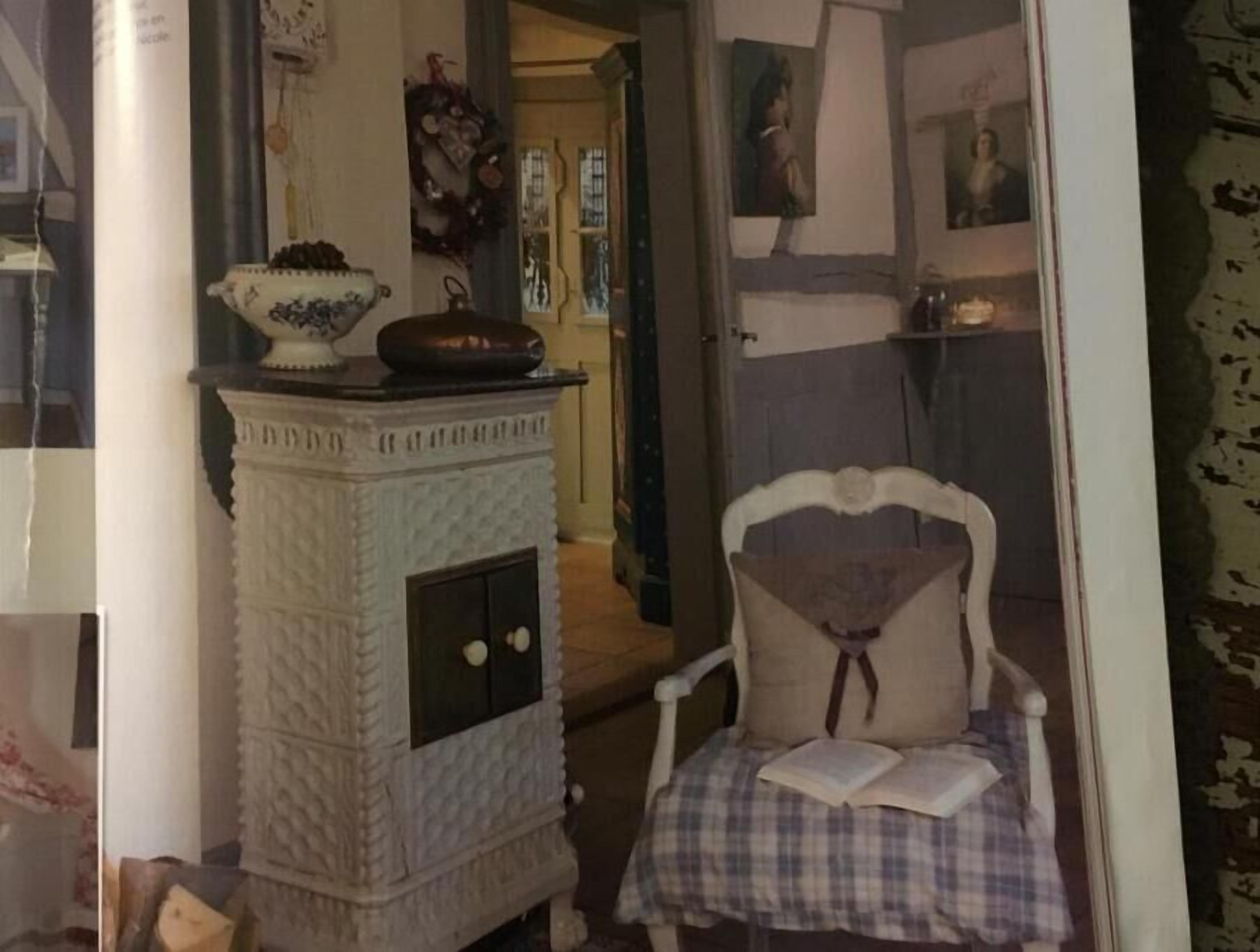 Une pièce avec une cheminée blanche à la texture intéressante, un fauteuil recouvert d’un tissu à carreaux et un livre posé dessus, ainsi que divers éléments décoratifs sur les murs.