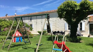 Un parque infantil con columpios y un tobogán, rodeado de un césped cuidado y un edificio de piedra con ventanas.