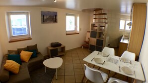 A compact living space with a sofa, a dining table with chairs, a small kitchen area, and a staircase.