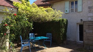 An outdoor patio with a table and chairs, surrounded by greenery and a house with a sloped roof.