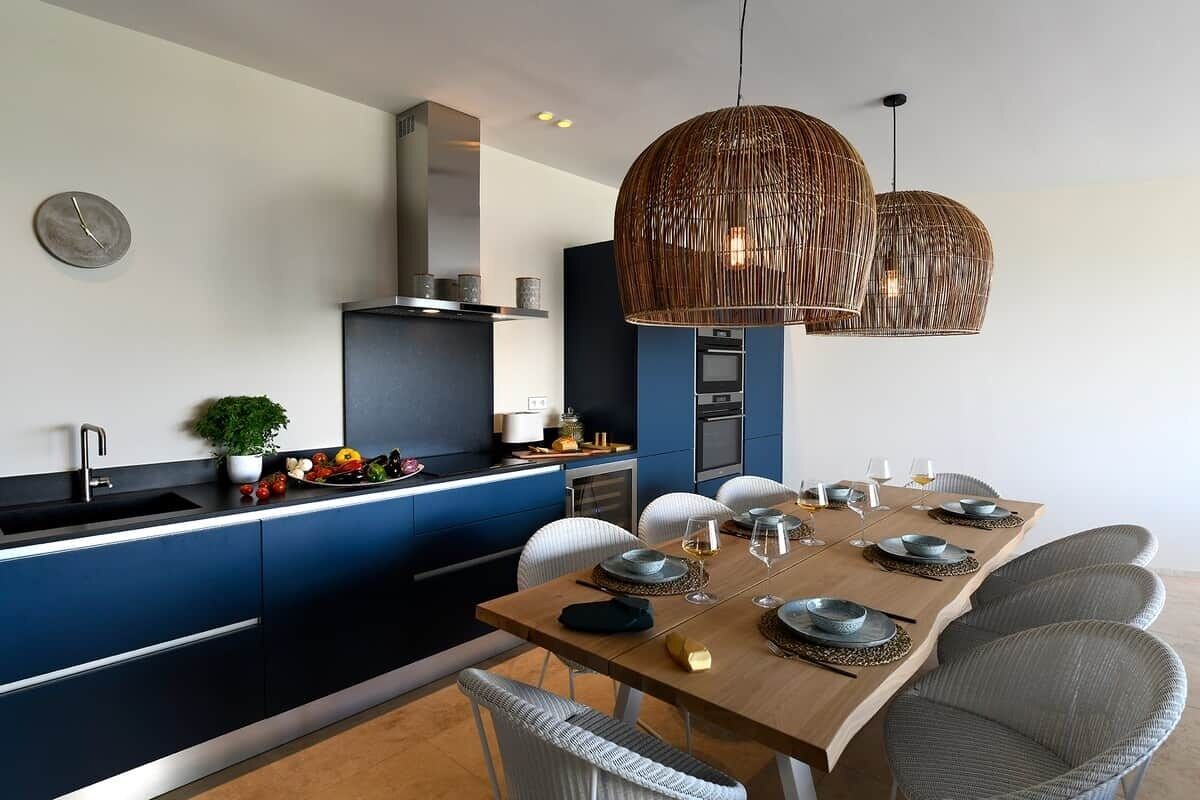 Une cuisine moderne avec des meubles bleus, un coin repas doté d’une table en bois pour six personnes, et deux grands luminaires suspendus en osier.