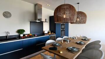 Une cuisine moderne avec des meubles bleus, un coin repas doté d’une table en bois pour six personnes, et deux grands luminaires suspendus en osier.