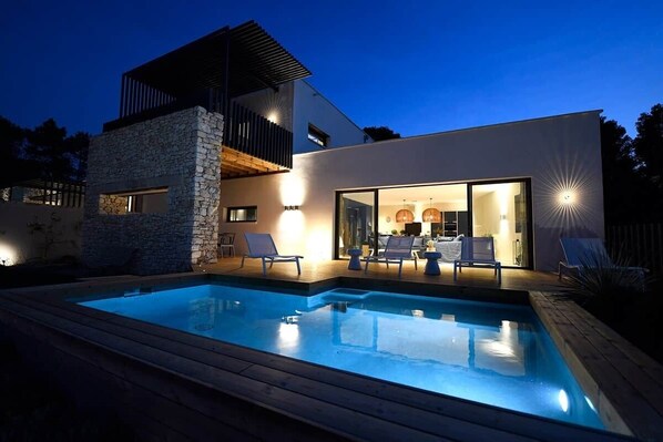A modern house with a swimming pool, outdoor seating, and a well-lit interior.