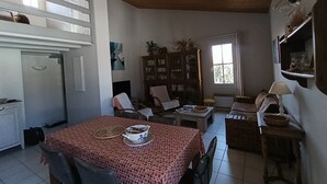A dining area with a table covered in a patterned tablecloth, surrounded by chairs. There is a bookshelf with books and decorative items, a window with natural light, and a wooden ceiling. - Holiday rental 5 people ideally located in St Martin de Ré (Saint-Martin-De-Ré)