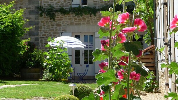 Um edifício de pedra com telhado de telhas, um jardim com flores cor-de-rosa e um guarda-sol branco.