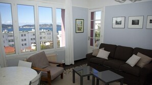 A living room with a brown sofa, a round table, and a view of buildings and the sea through large windows. - For six people, 1st floor of villa, sea view apartment of 80m2 (Toulon)