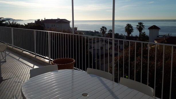 A balcony with a table and chairs, overlooking a coastal view with houses and palm trees. - For six people, 1st floor of villa, sea view apartment of 80m2 (Toulon)