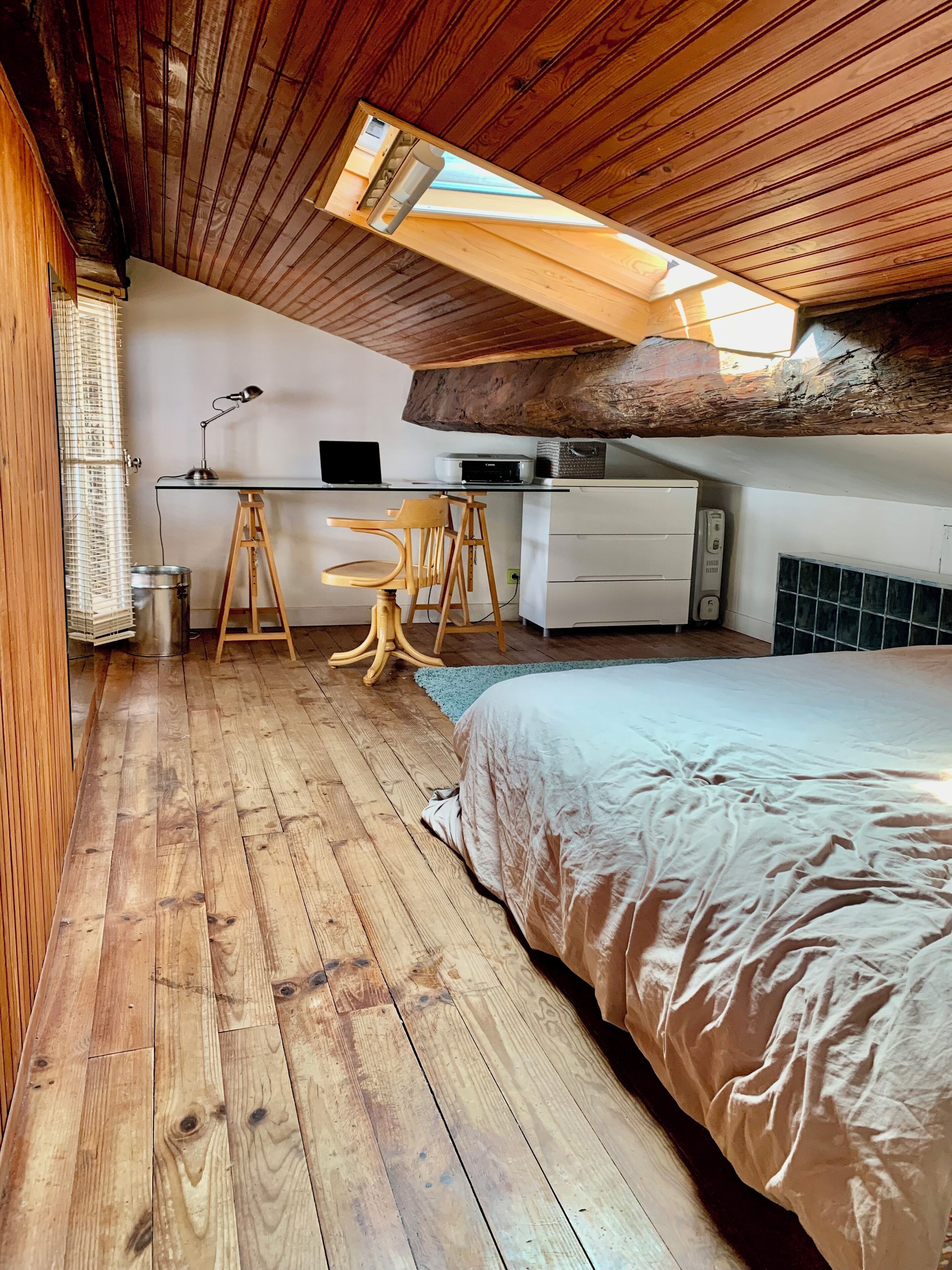 A loft bedroom with a wooden floor, a bed, a desk with a chair, a computer, and a radiator.