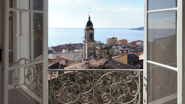 Una ventana abierta con vistas a un paisaje urbano costero, que incluye una torre destacada y edificios con tejados de tejas rojas.