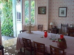 Uma sala de jantar com uma mesa posta para quatro pessoas, rodeada de cadeiras. Há um candelabro sobre a mesa e uma janela com vista para o verde lá fora.