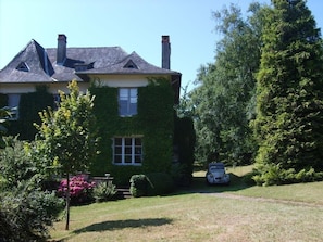 Uma casa com telhado íngreme, várias chaminés e um jardim bem cuidado com gramado e diversos arbustos.