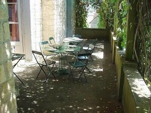 Um pátio ao ar livre com uma mesa comprida e cadeiras, rodeado por paredes de pedra e vegetação.