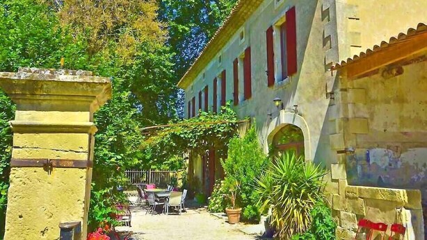 Un patio con un edificio que tiene contraventanas rojas, una pared de piedra y plantas en macetas.