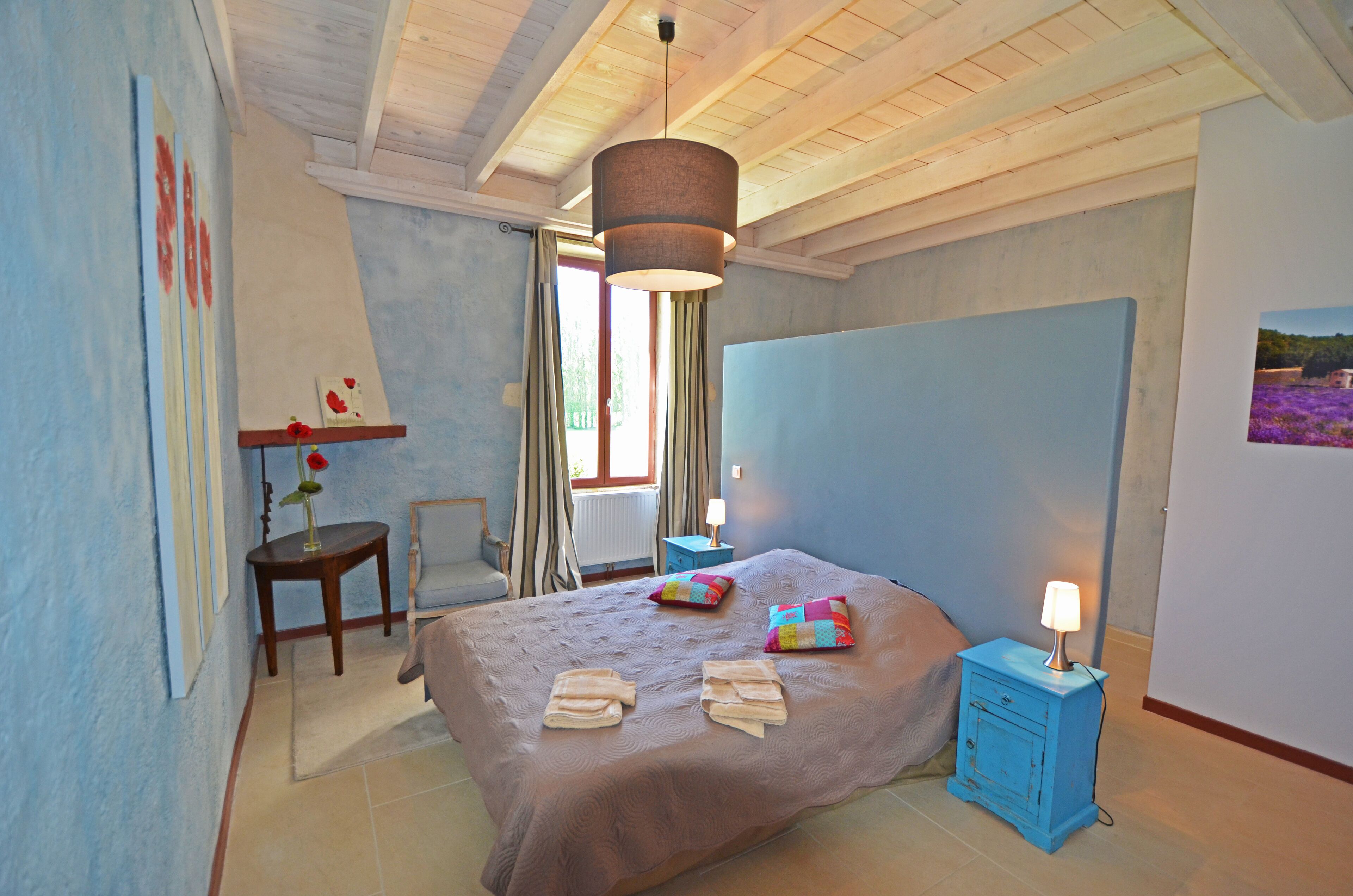 A bedroom with a large bed, a bedside table with a lamp, a chair, and a window with curtains.