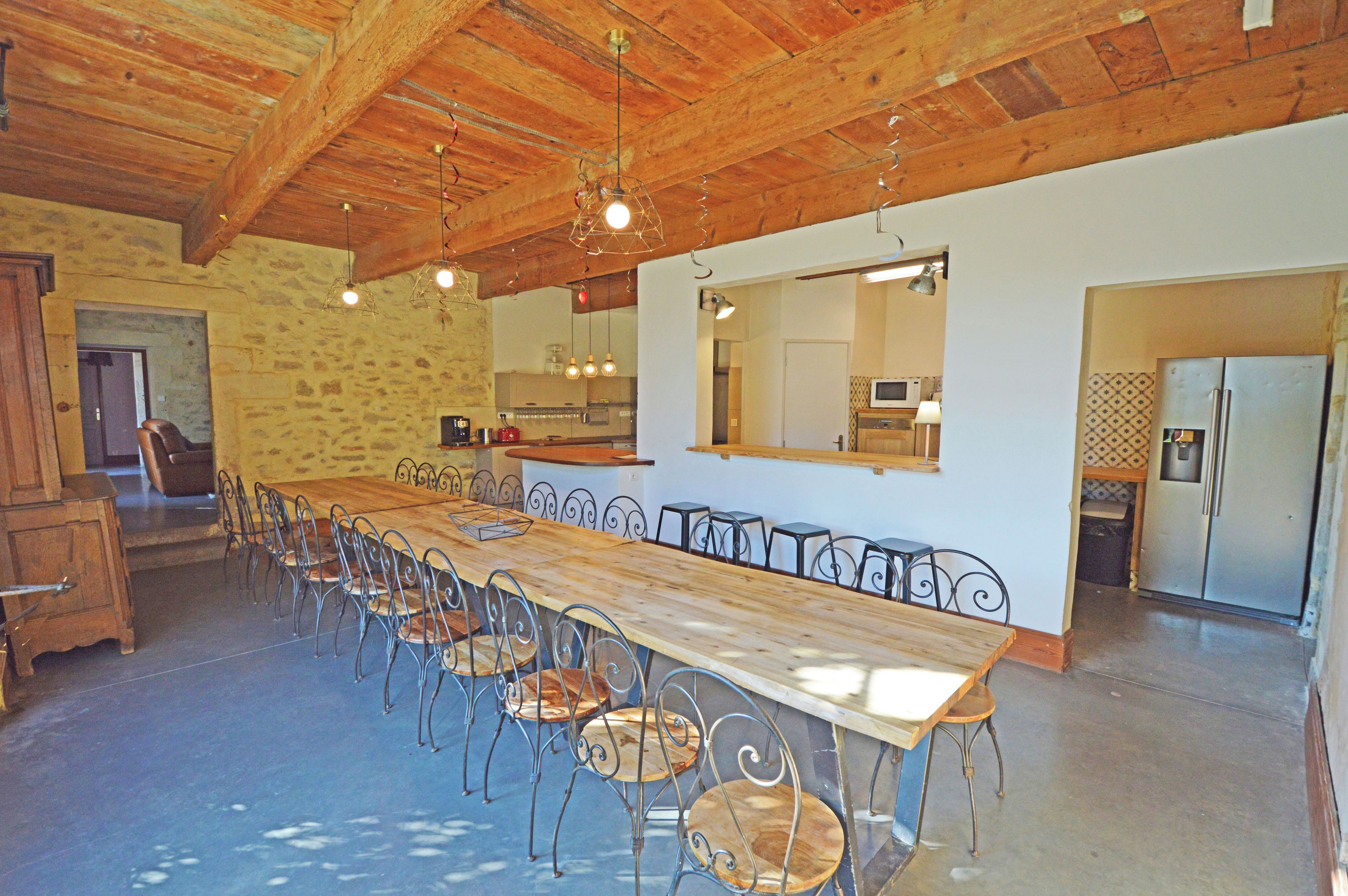 Un grand espace à manger avec une longue table en bois et des chaises en métal, une cuisine en arrière-plan et un mur en pierre.