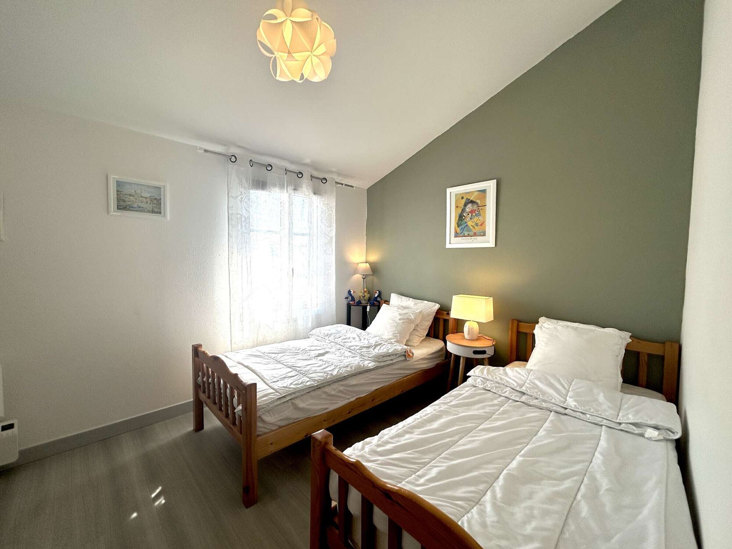 Une chambre d’hôtel avec deux lits simples, une table de chevet avec une lampe, une fenêtre avec des rideaux et un tableau accroché au mur.