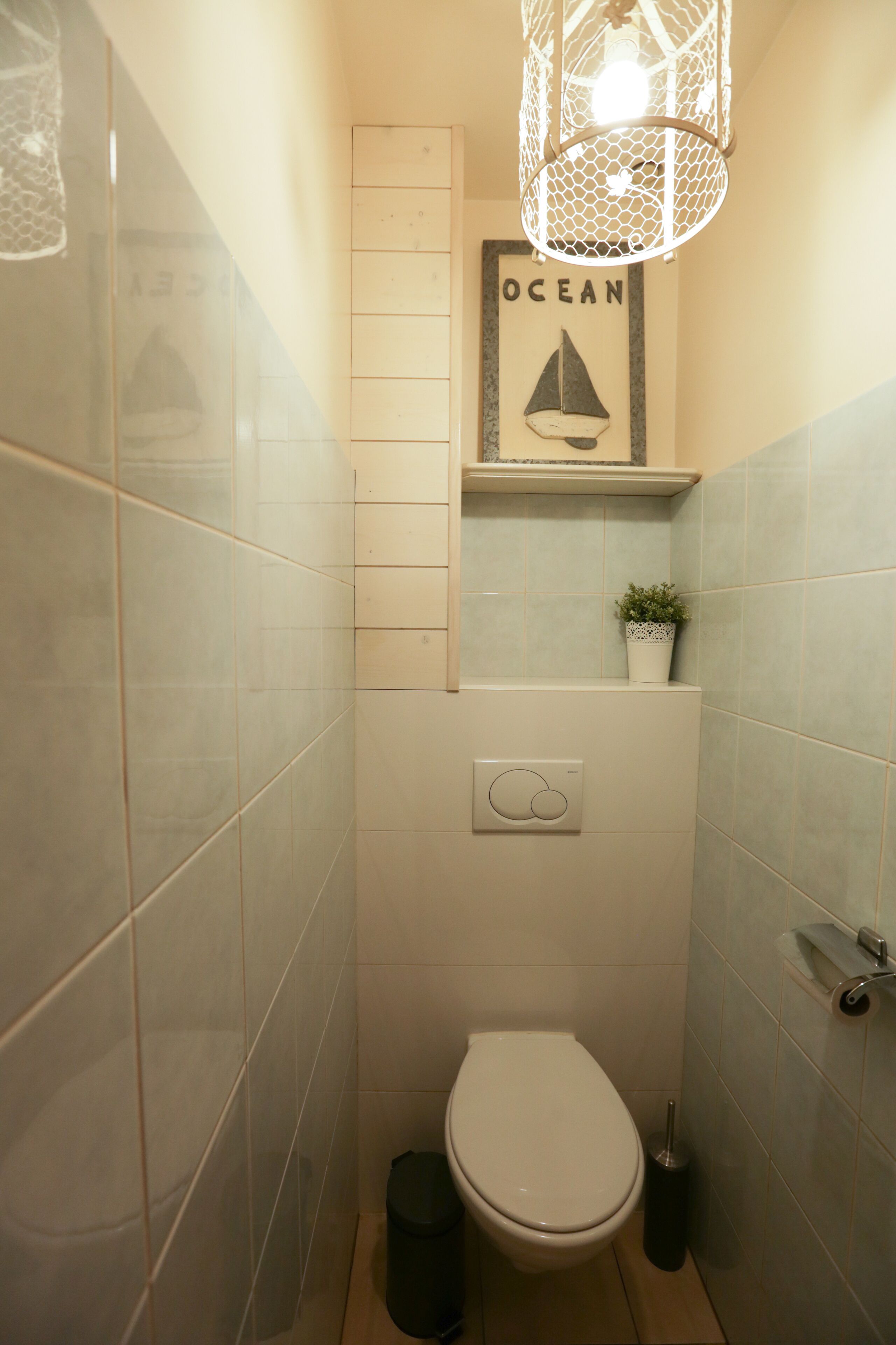A small bathroom with a toilet, a trash bin, a wall-mounted shelf with a potted plant, and a framed picture of a sailboat with the word "OCEAN" above it.