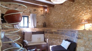 A stone-walled room with a bed, a black armchair, a small table with a vase, and a hanging light fixture.