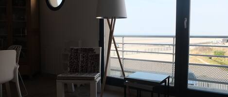 Una habitació amb una finestra gran, una cadira blanca, una cadira amb dibuixos, una taula petita, una làmpada i una prestatgeria.