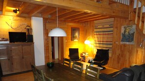 Ein Holzinterieur mit einem Esstisch, Stühlen, einem schwarzen Ledersofa, einem Fernseher und einer Lampe.