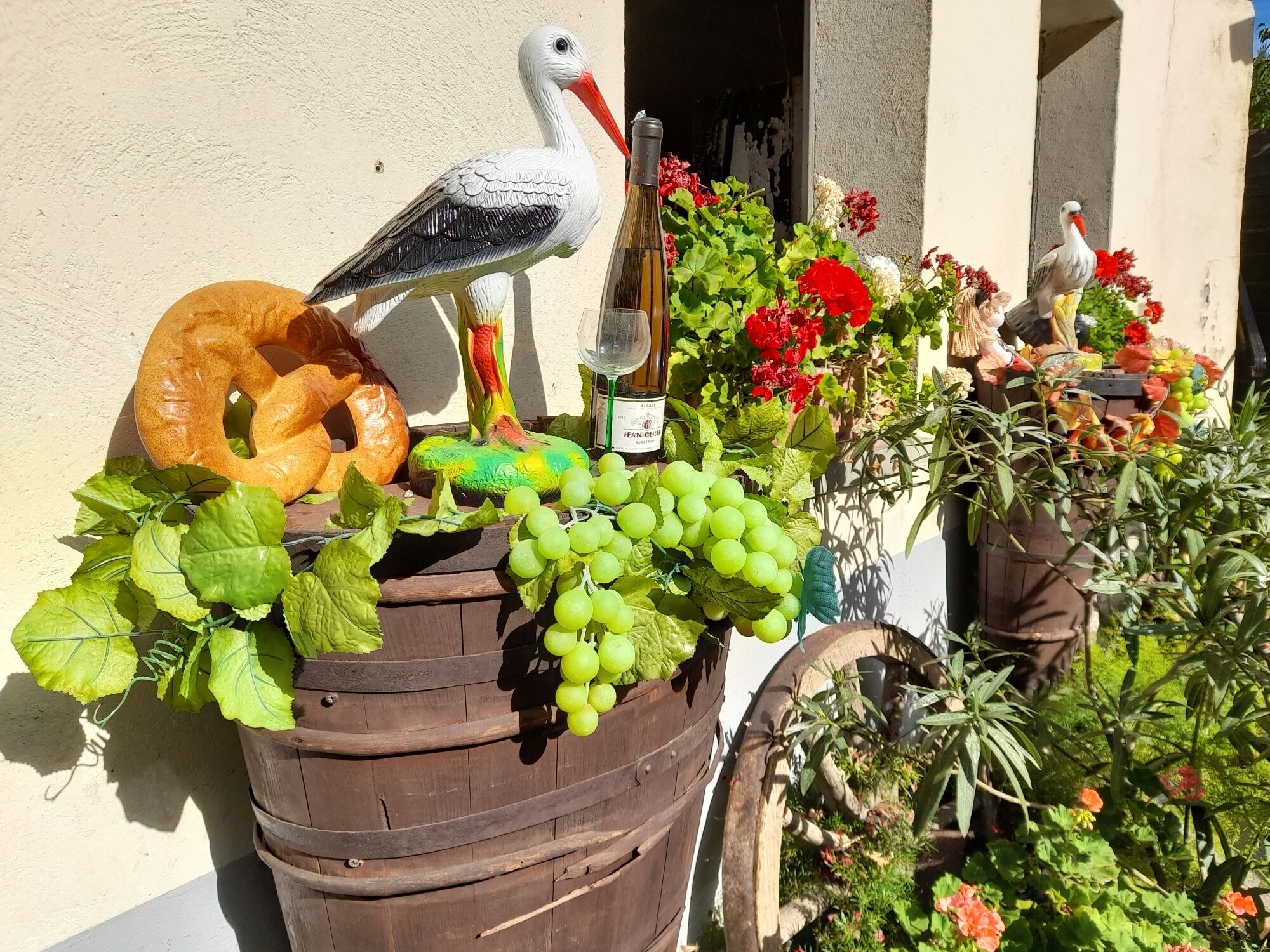 A wooden barrel filled with green grapes, a loaf of bread, and a bottle of wine, surrounded by colorful flowers and a bird statue.