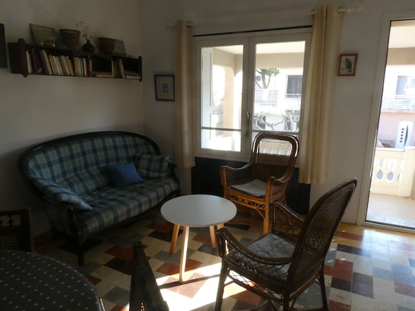 A living room with a checkered sofa, a wicker chair, a small white table, and a bookshelf.