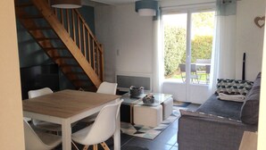 Uma sala de jantar com mesa de madeira e cadeiras brancas, escada, sofá com almofadas estampadas e vista para o exterior por uma porta de vidro.