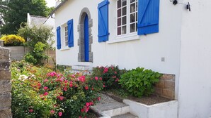 Ein weißes Gebäude mit blauen Fensterläden und einem steinernen Bogen. Treppen führen zum Eingang, und ein Garten mit rosa und weißen Blumen ist angelegt.