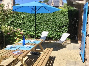 Ein Holztisch mit zwei Stühlen, einem blauen Sonnenschirm und einem Garten mit dichtem Grün im Hintergrund.