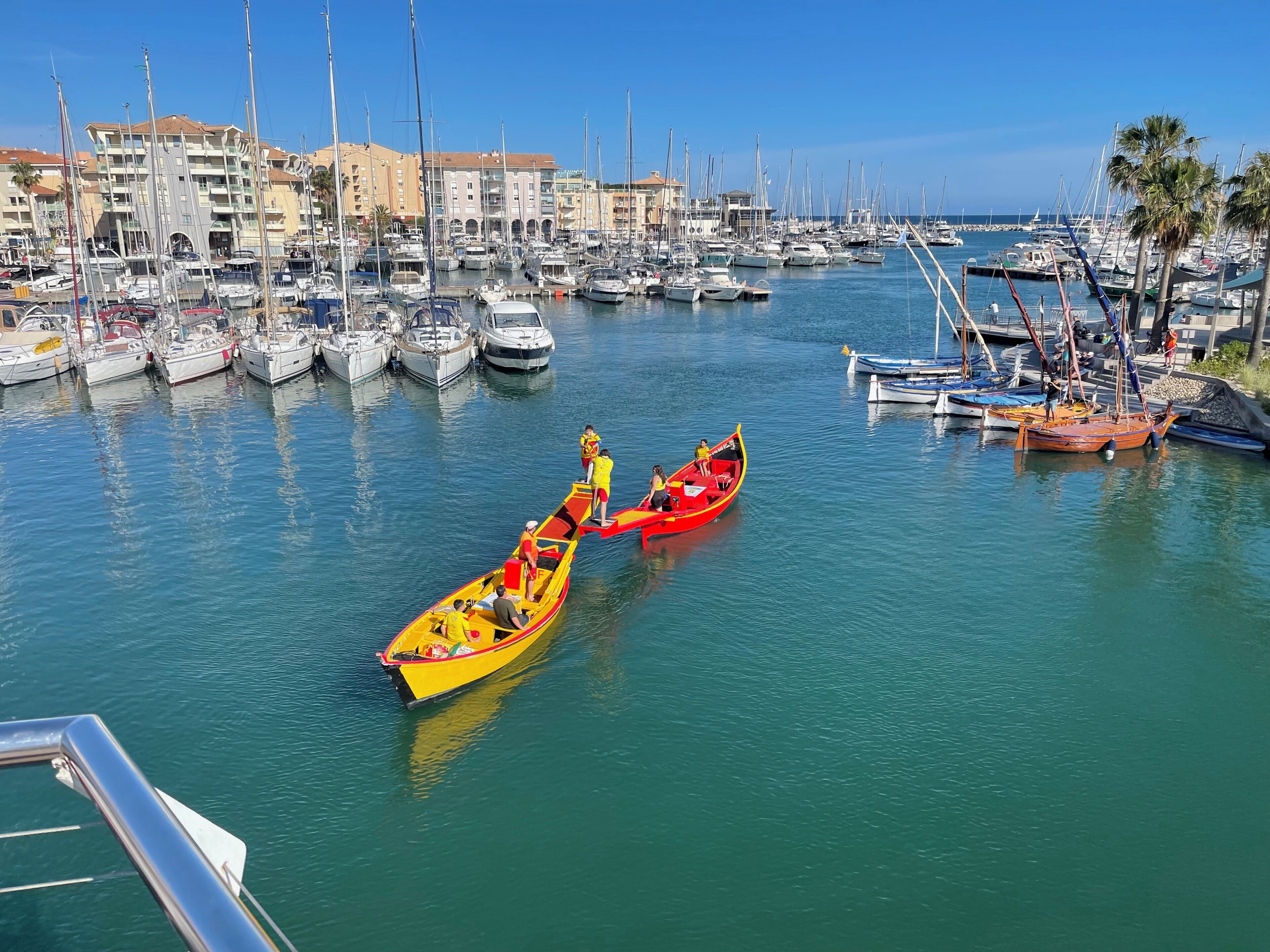 A marina with multiple boats docked, a red boat with passengers, and buildings in the background.