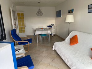 Una stanza con un divano bianco, una poltrona blu, un tavolo da pranzo con sedie, una lampada e una porta con un oblò giallo.