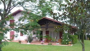 Une maison avec un toit rouge et des murs blancs, entourée d’une pelouse bien entretenue et de différentes plantes en pot.