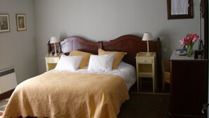Un lit bien fait, avec une tête de lit en bois, deux tables de chevet avec des lampes, un bureau agrémenté d’un vase de fleurs et un tableau encadré accroché au mur.