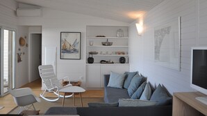 A modern living room with a grey sofa, white rocking chairs, and a small table. There are shelves with decorative items and a framed picture on the wall. - MAGNIFICENT, COMFORTABLE TRADITIONAL CHALET FACING THE SEA (Gruissan)