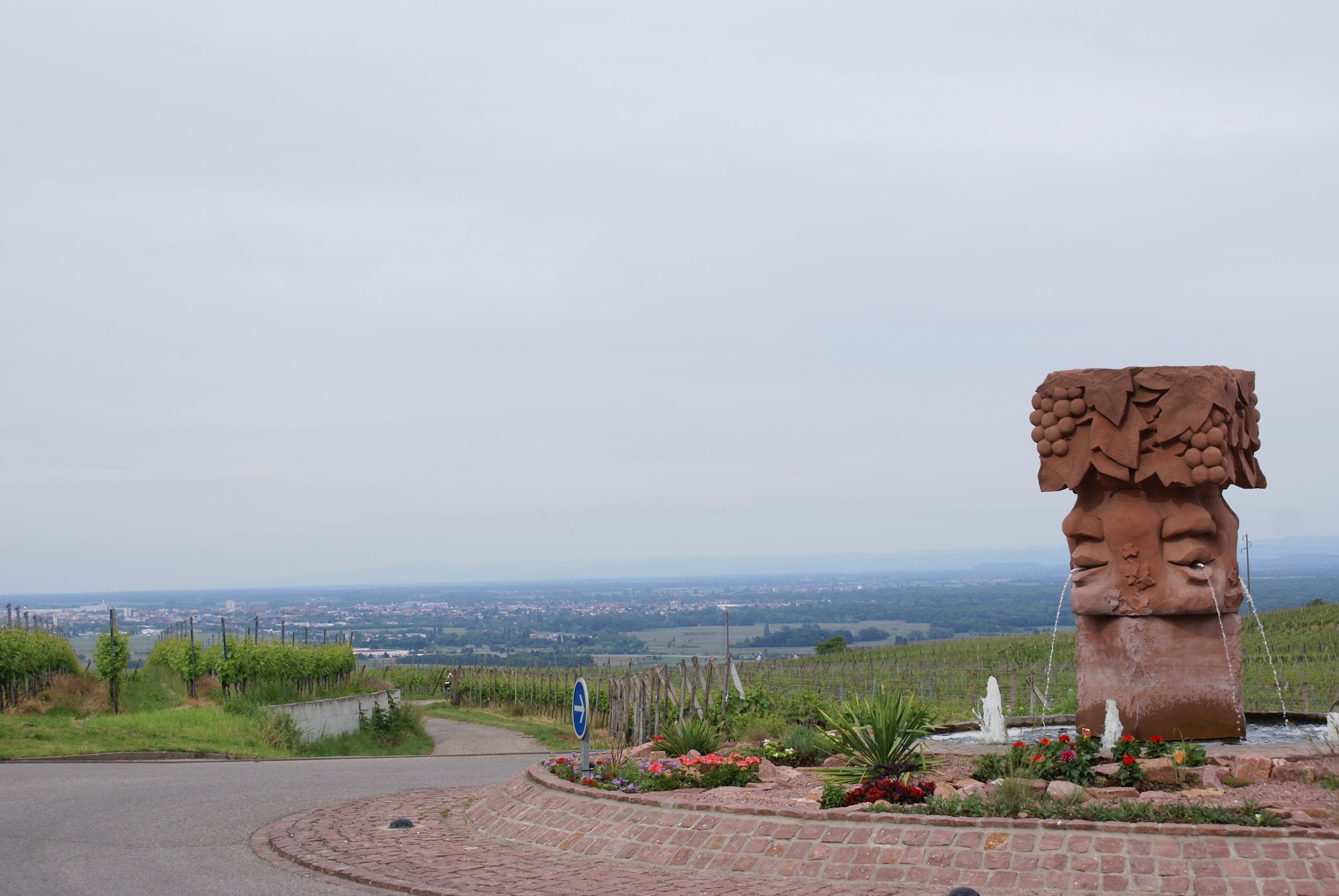 A stone sculpture with a grapevine design, surrounded by a landscaped garden with flowers and a view of a distant cityscape.