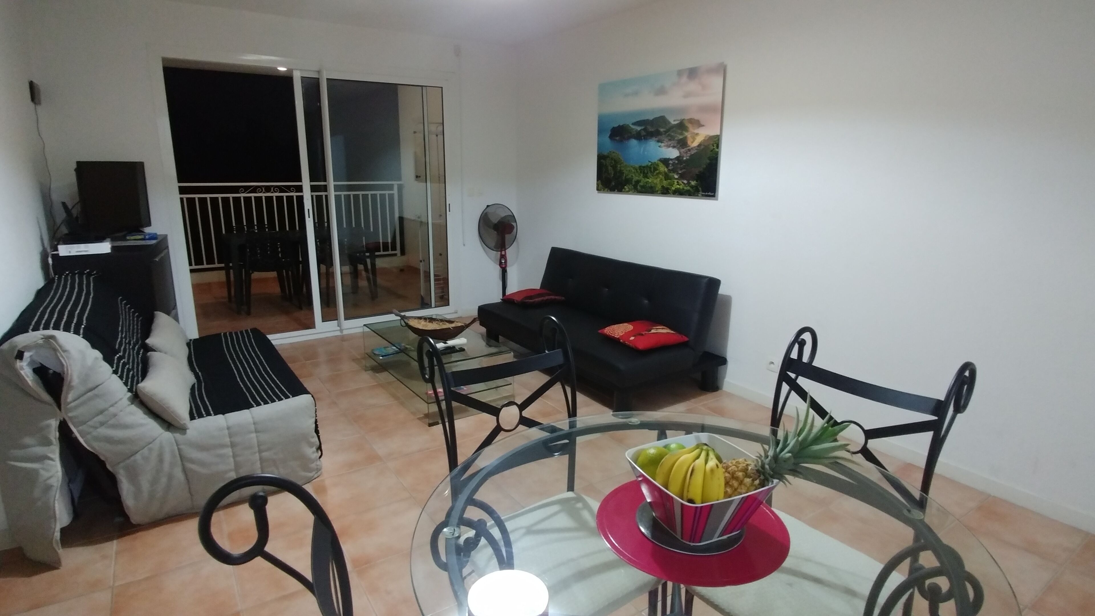 A living room with a sofa, a glass table with fruit, a fan, and a sliding glass door leading to an outdoor area.