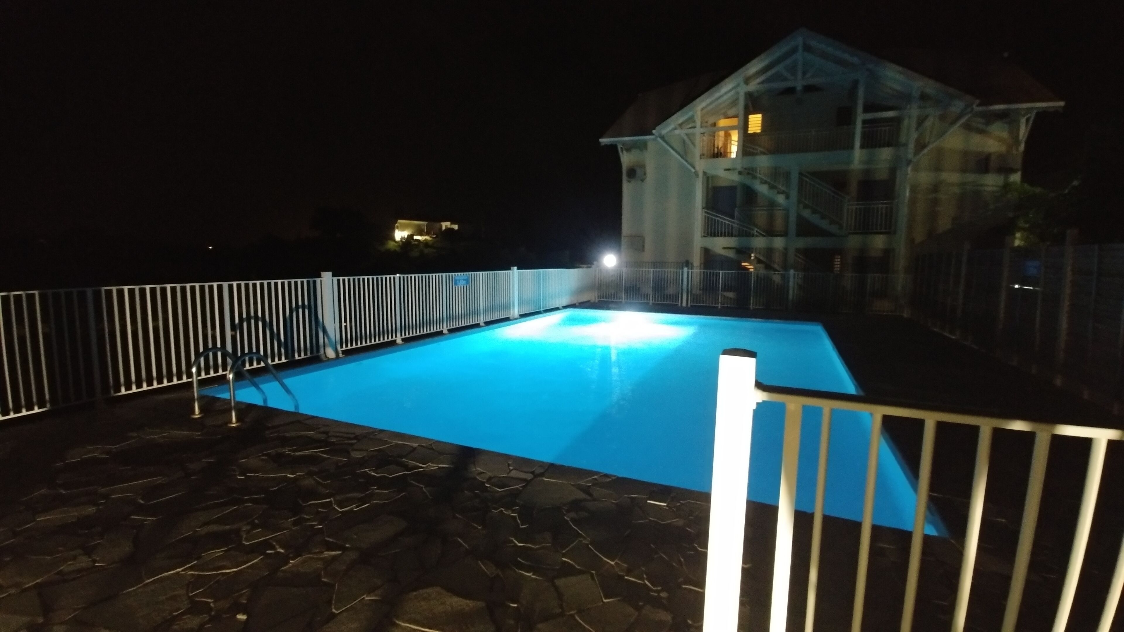 An outdoor swimming pool with a stone deck, surrounded by a white fence, and a building with a balcony in the background.