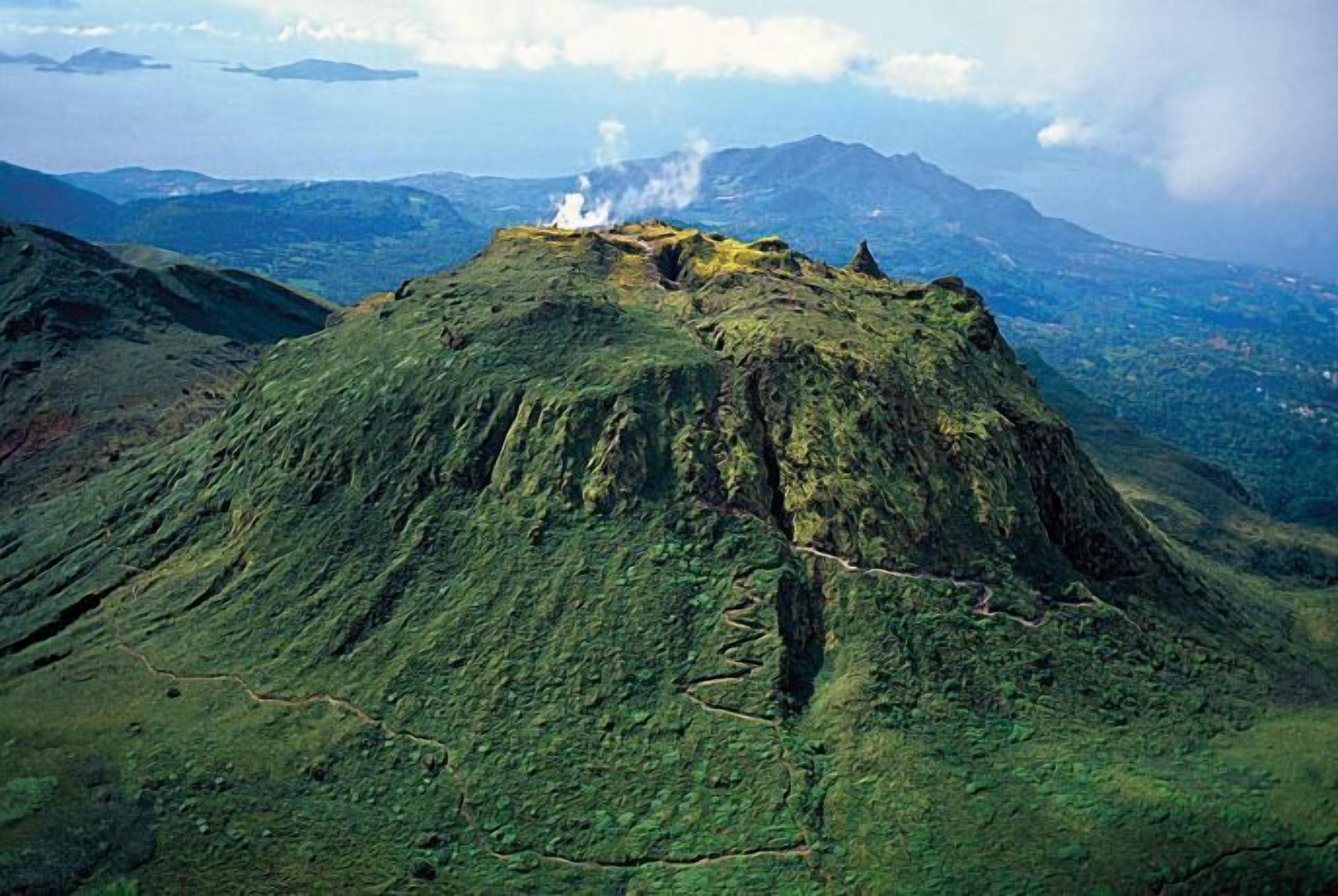 A volcanic landscape with a prominent peak emitting smoke, surrounded by lush greenery and distant mountains.
