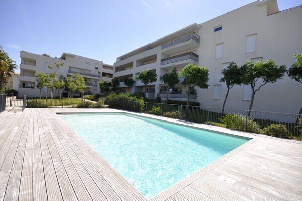 A swimming pool with a wooden deck, surrounded by modern buildings with balconies and trees.
