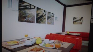 Een ontbijtruimte met een tafel gedekt, inclusief schalen met fruit, gebak en drankjes. Er zijn rode bekleden zitjes en een witte muur met abstracte kunst.