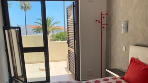 Een slaapkamer met een bed overdekt met een stippenbedspreide, een rood kussen en twee opgerolde handdoeken. Er is een schuifdeur die leidt naar een buitenruimte met uitzicht op palmbomen en gebouwen.