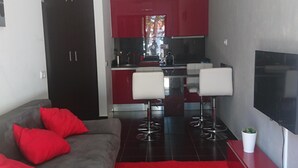 Een moderne woonkamer met een grijze bank, rode kussens, een rood vloerkleed, een televisie en een keuken op de achtergrond.