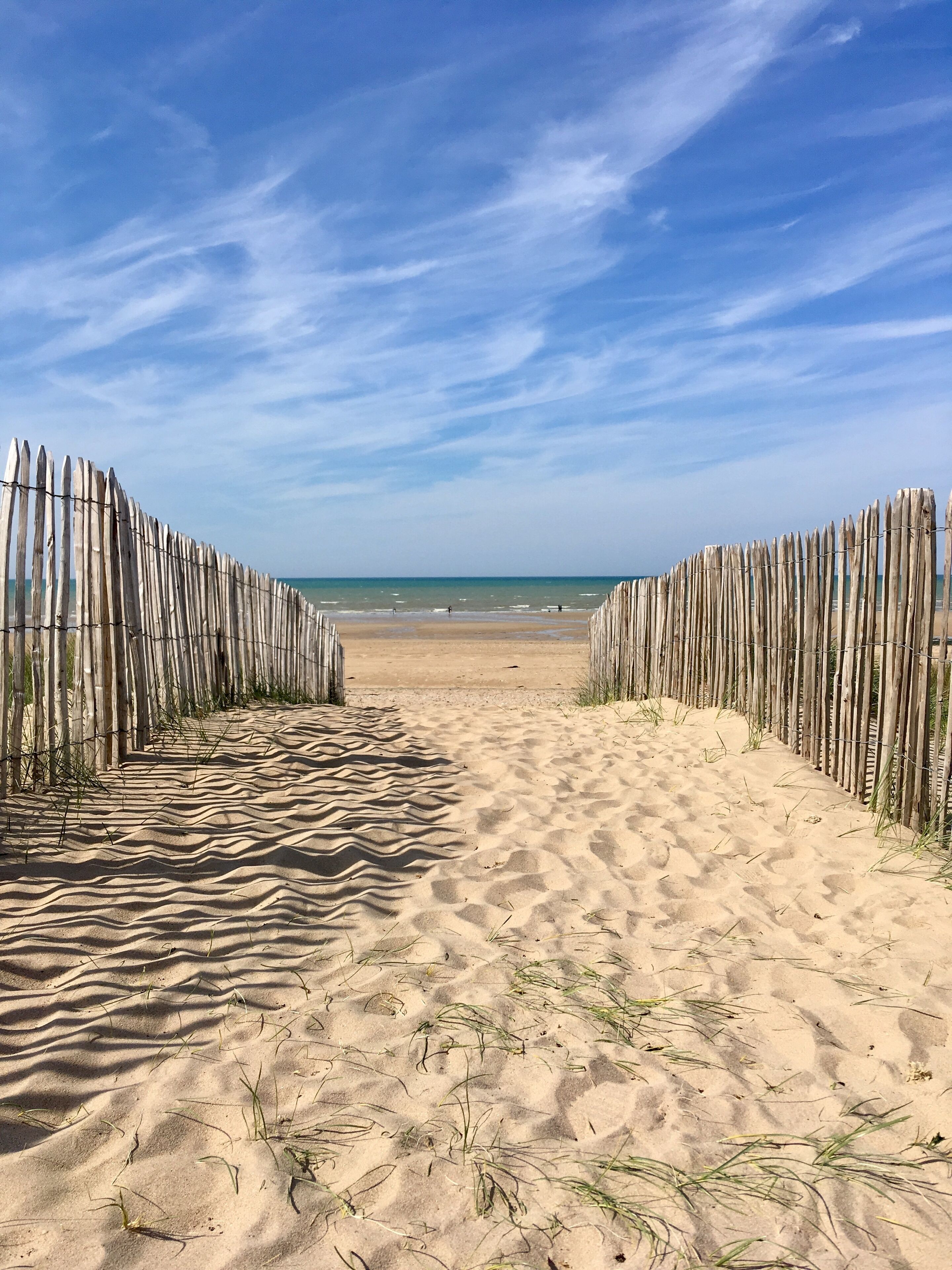 Un sentier de sable qui longe la plage et mène à l’océan, bordé de clôtures en bois.