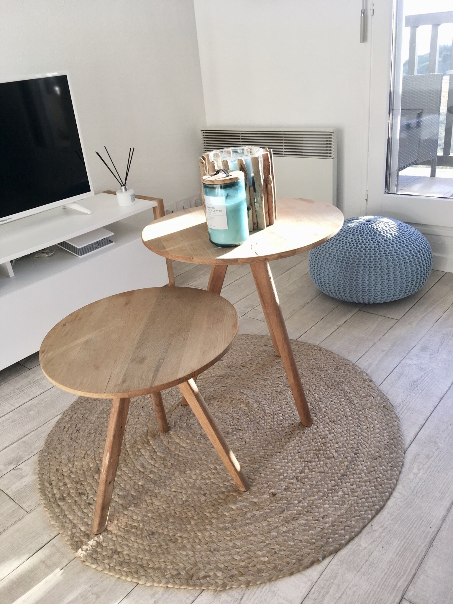 A modern living room with a TV, two wooden tables, a blue candle jar, and a woven pouf.