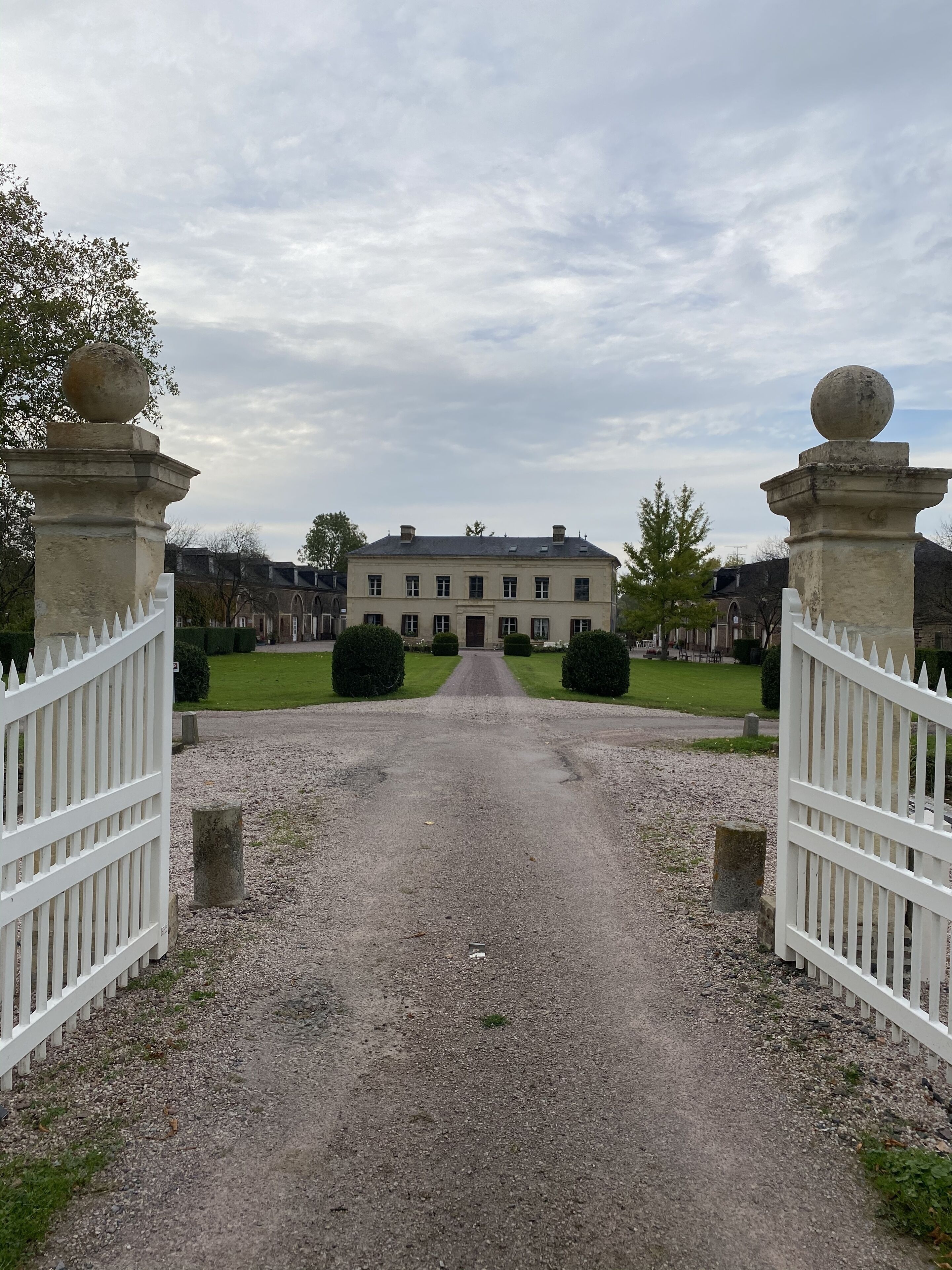 A gravel driveway leading to a large, symmetrical building with a central entrance, flanked by white picket fences and stone bollards.