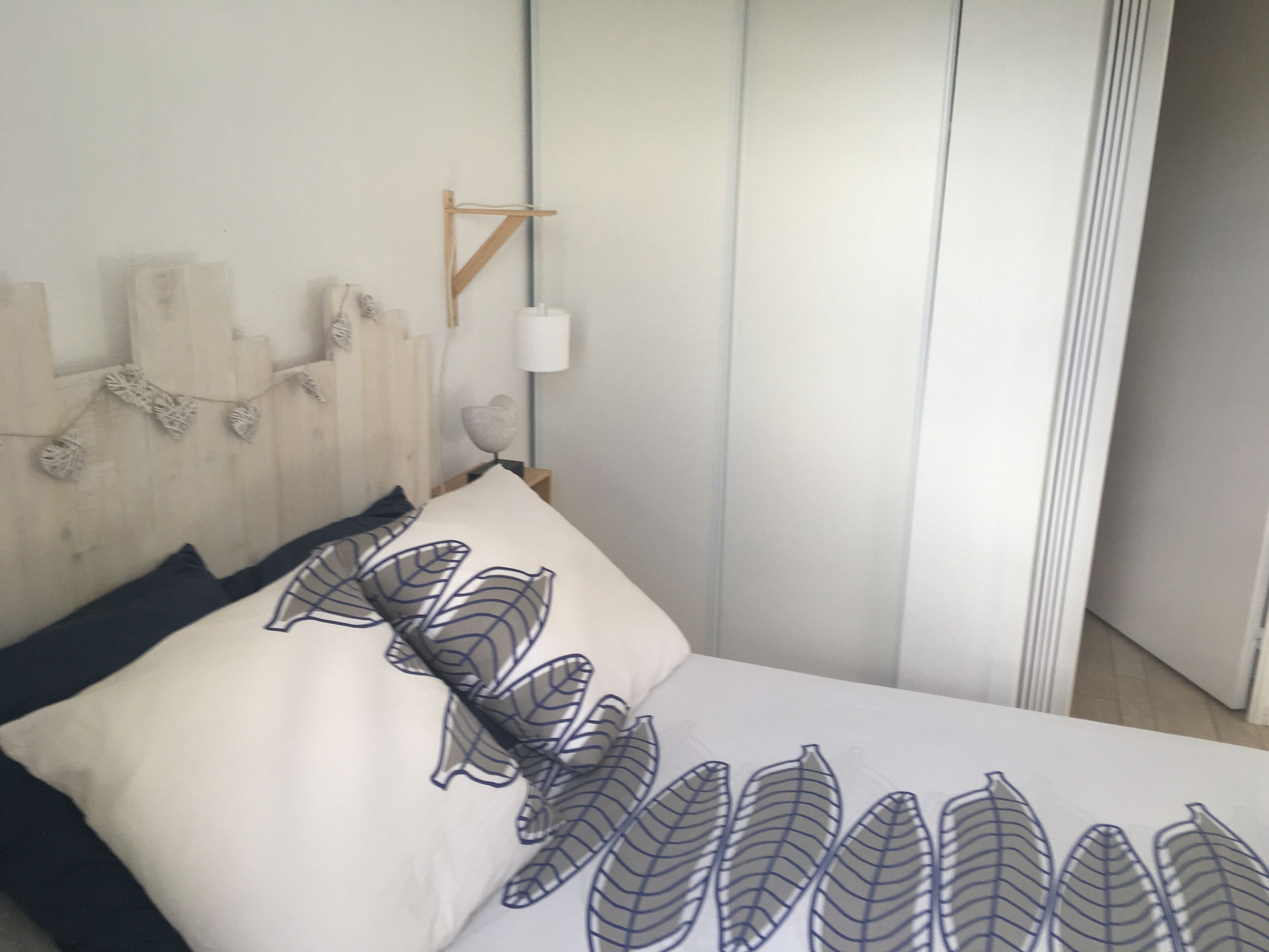 A bedroom with a wooden headboard, a bed with a white and black leaf-patterned bedspread, and a bedside table with a lamp.