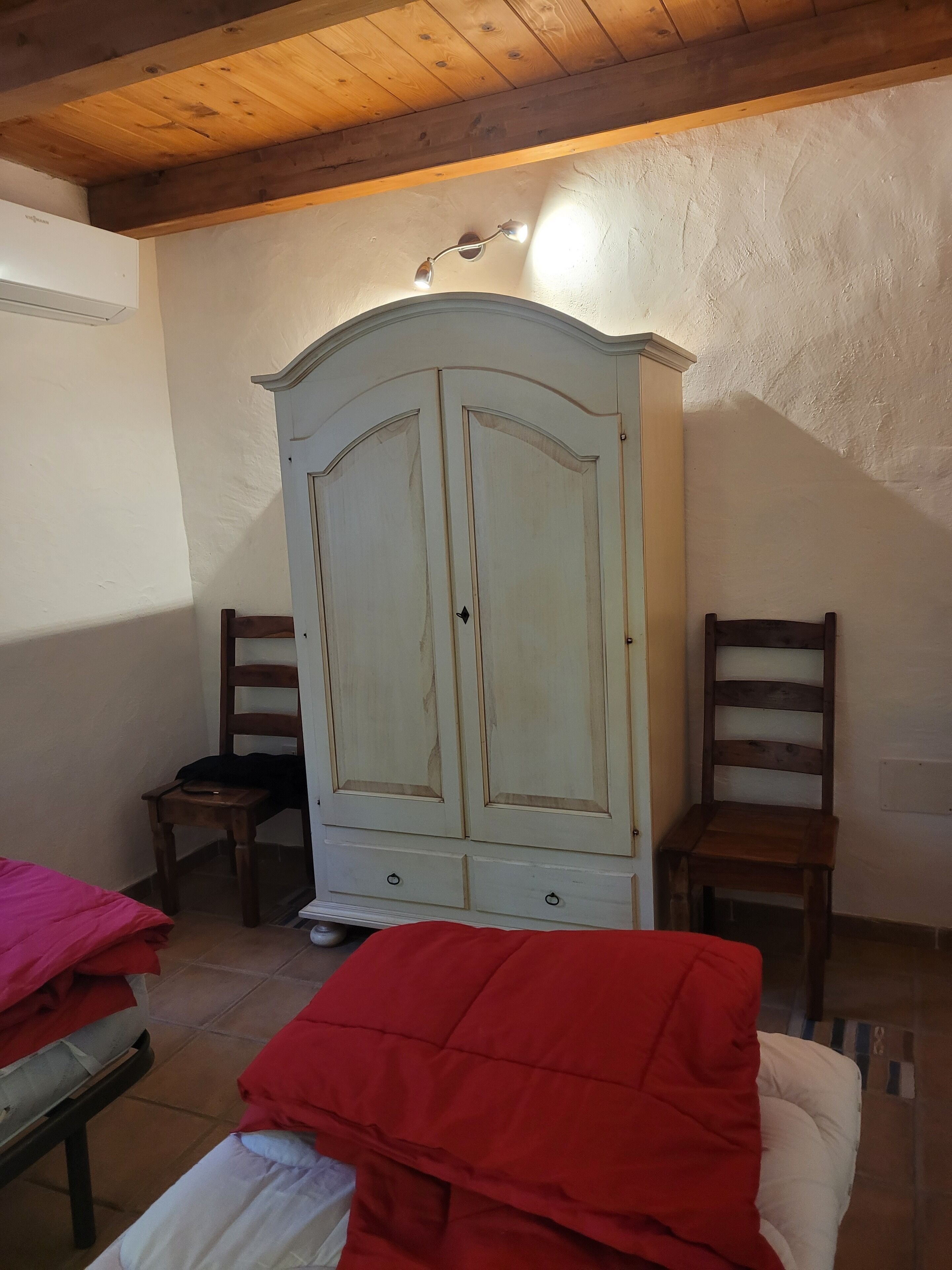 A room with a wooden ceiling, a white wardrobe with two doors, two wooden chairs, a bed with a red blanket, and a small table with a chair.