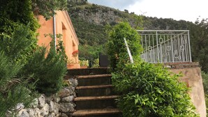 A stone staircase leading up to a building with a metal gate, surrounded by greenery and a mountainous backdrop. - Small villa located in a quiet pool and sea view / Gulf of Policastro (MARATEA)