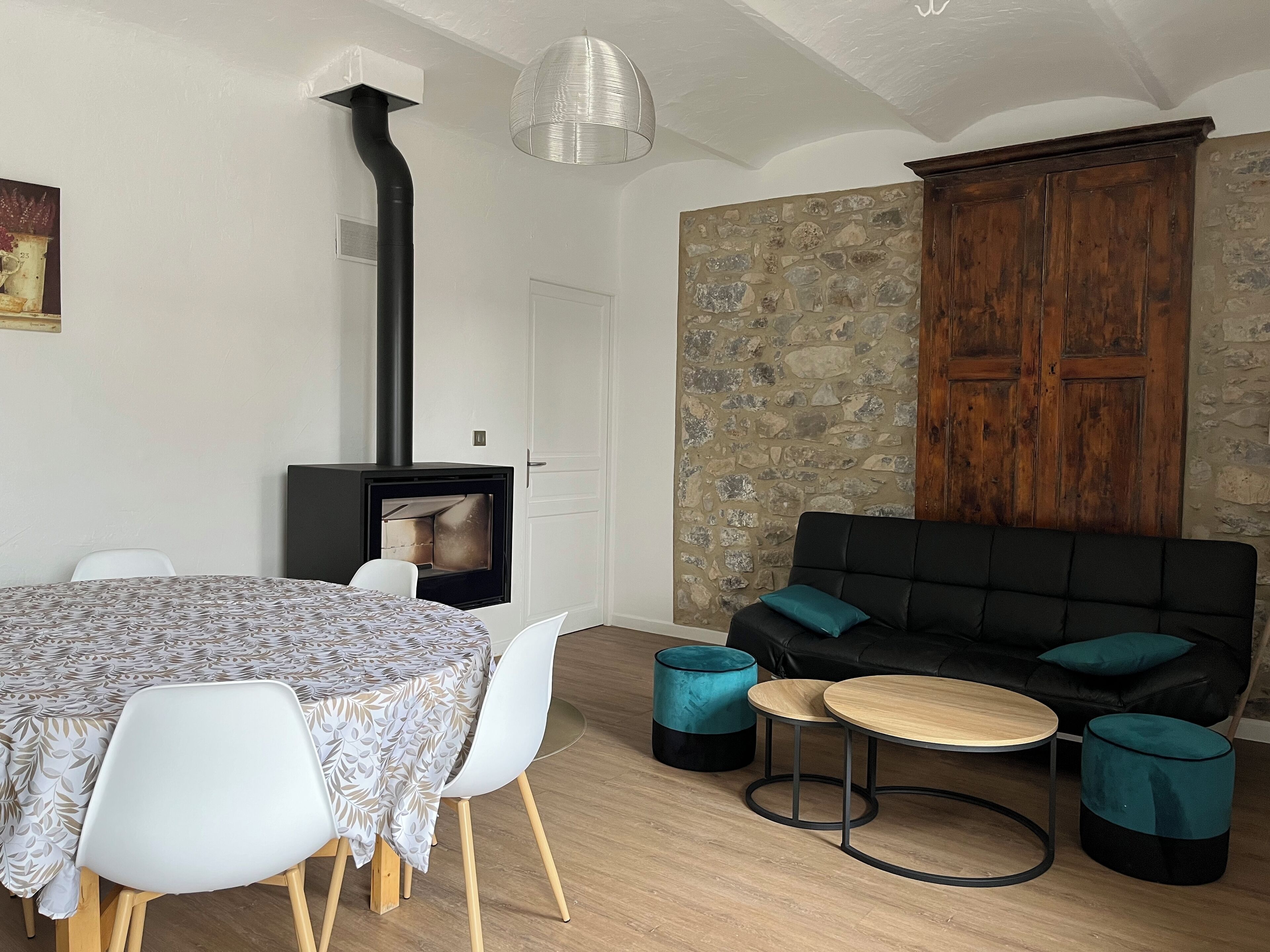 Una sala acogedora con una pared de piedra, un sofá negro con cojines color turquesa, una mesa de centro redonda de madera y una mesa de comedor blanca con sillas.